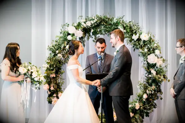 Couple holding hands and exchanging vows in front of white-and-green circle arch