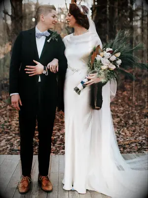 couple standing outside in front of forest with bride holding pageant style wedding bouquet of winter evergreens and roses