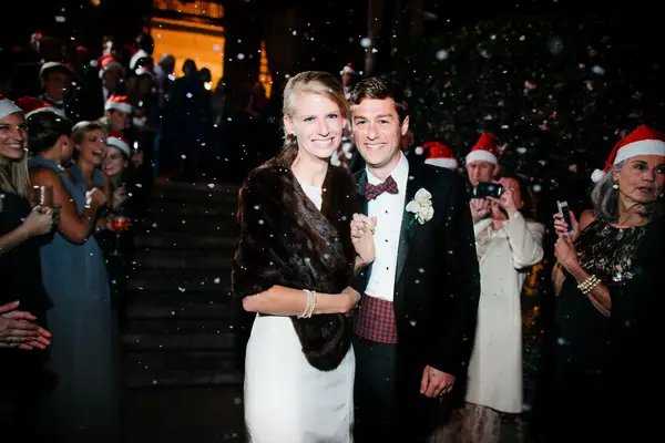 newlyweds exit wedding reception with fake snow and guests wearing santa hats