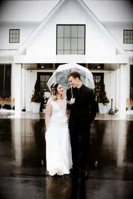 bride and groom walk underneath a clear umbrella outside of rustic wedding venue