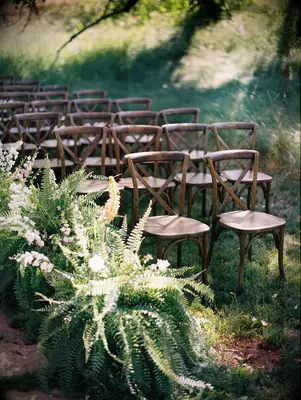 outdoor wedding ceremony aisle lined with large sword fern plants next to wooden crossback chairs