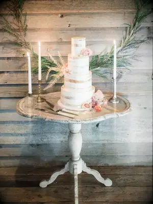 modern three tier semi naked wedding cake on whitewashed wooden table decorated with blush roses and bleached greenery