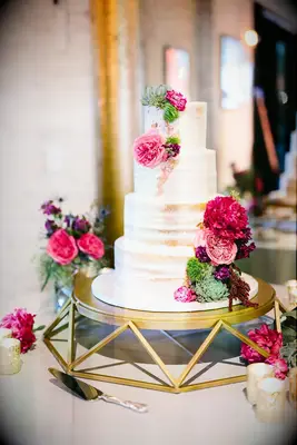 four tier semi naked wedding cake with bright pink roses, dahlias and succulents on geometric gold cake stand