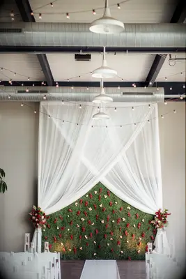 modern wedding ceremony backdrop at industrial venue with white draping, greenery wall, red flowers, bistro lights across the ceiling and white chairs