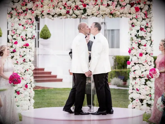 two grooms kiss in front of pink and white square flower backdrop at outdoor wedding ceremony
