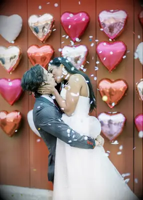groom kisses bride as he picks her up in front of a wall decorated with pink, red and silver heart-shaped balloons