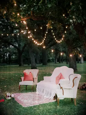 outdoor wedding lounge area with white vintage loveseat and armchairs, pink throw pillows, pink rug and twinkle lights in the trees