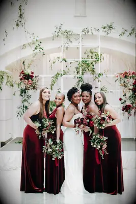 bride poses with bridesmaids wearing red velvet floor length bridesmaid dresses