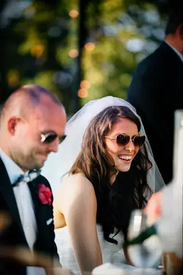 candid smiling photo of bride wearing heart-shaped sunglasses and veil sits next to groom at outdoor wedding reception table