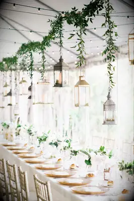 long wedding reception table with greenery garland and mismatched silver and gold lanterns hanging above