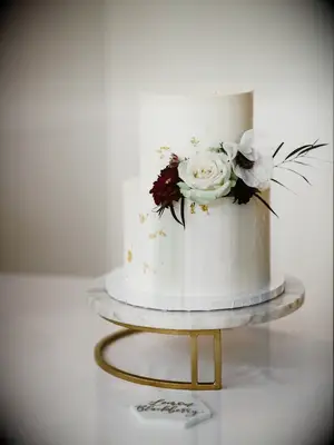 white two tier buttercream wedding cake decorated with edible gold leaf and burgundy, light green and white flowers on white marble cake stand