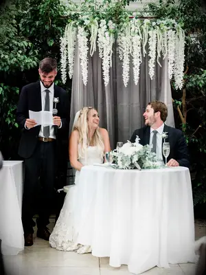 Best man giving toast at wedding reception while bride and groom laugh