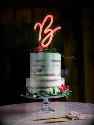 Rustic two-tier wedding cake with greenery and neon initial cake topper