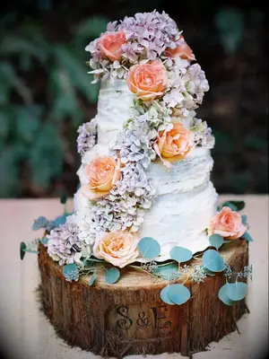 Three-tier rustic wedding cake with hydrangeas and roses on custom wood slice cake stand with initials