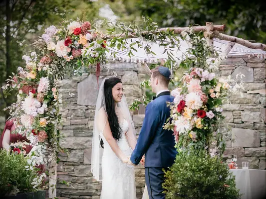 bride and groom standing under chuppah made from birch tree branches decorated with flowers and greenery