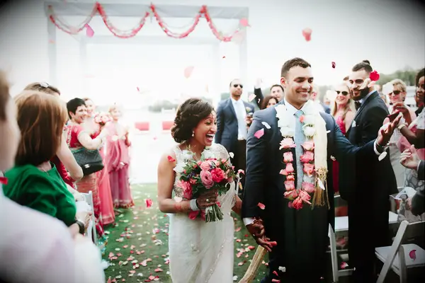 bride and groom hold hands and smile as guests toss petals into the wedding aisle
