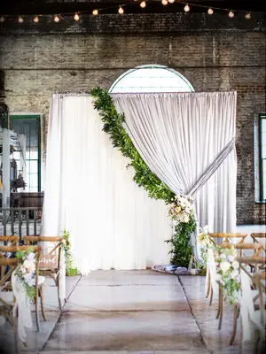 indoor wedding ceremony backdrop with curtain draped to one side decorated with greenery garland and flower tie-back