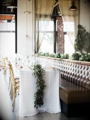 long wedding banquet table with white tablecloth, greenery table runner and wooden cross-back chairs