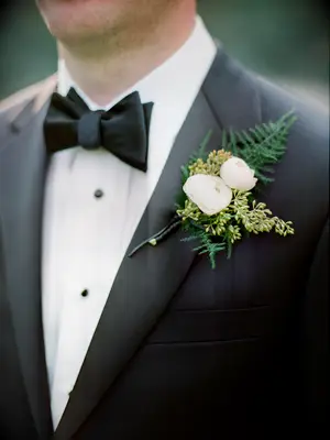 white ranunculus wedding boutonniere with greenery pinned to black tuxedo lapel