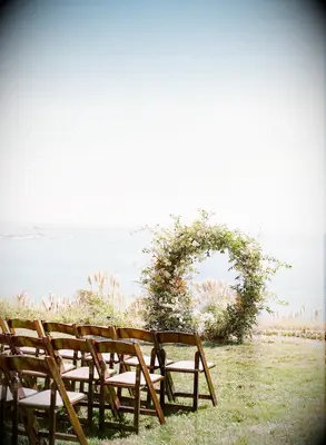 Waterfront wedding ceremony with wood folding chairs and greenery full-circle arch