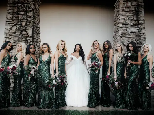 bride stands against a wall with bridesmaids wearing floor-length sequin emerald green gowns