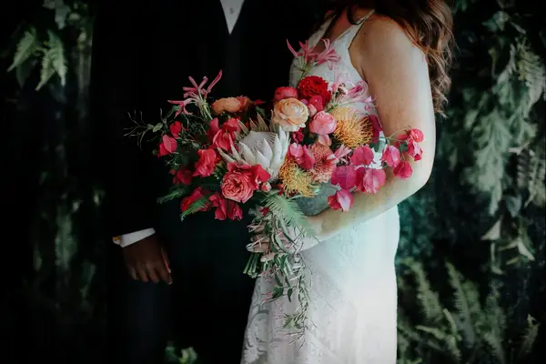Bride and groom hold a vibrant pink bouquet featuring dahlias, roses, and greenery. 