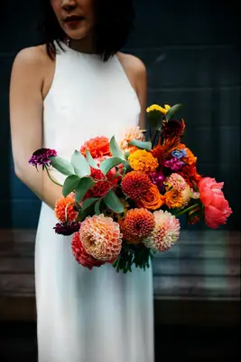 Bride holds a modern bouquet of orange, red, and blush pink dahlias. 