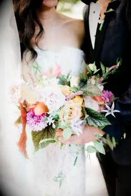 Couple holds a boquet of pastel-colored flowers, including dahlias, roses, and greenery. 
