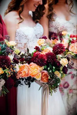 Bride and bridesmaids hold vibrant bouquets of dahlias and roses in red and orange hues. 