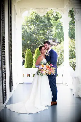 Bride and groom embrace while holding a beautiful bouquet featuring dahlias and other fresh flowers. 