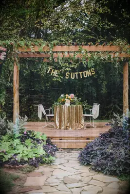 Outdoor sweetheart table under a wooden pergola with a banner hanging above that says the suttons