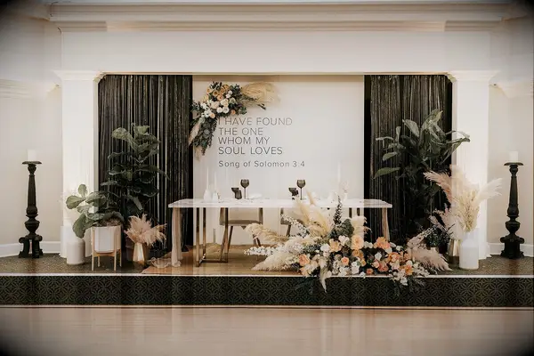 indoor wedding sweetheart table idea with table on a raised platform decorated with pampas grass arrangements, potted plants and a backdrop with bible verse from song of solomons 3.4