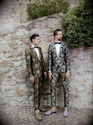 two grooms stand against a stone wall wearing metallic silver and gold brocade pattern tuxedos