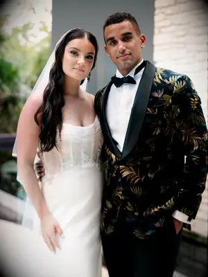 bride and groom pose for portrait wearing luxury wedding attire. bride is wearing strapless fit and flare gown with sheer corset bodice and groom is wearing black velvet tuxedo with tropical greenery print