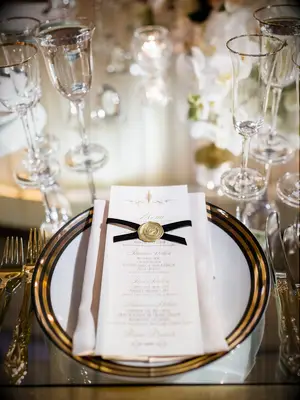 wedding menu card decorated with black ribbon and gold wax seal on top of gold-rimmed dinner plate