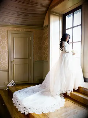 bride wearing lace ball gown wedding dress with cathedral train stands in front of floor length window holding her bouquet