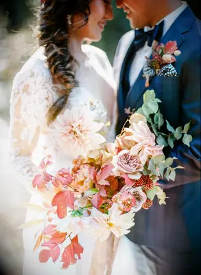 Bride and groom hugging and holding bouquet with leaves and dahlias