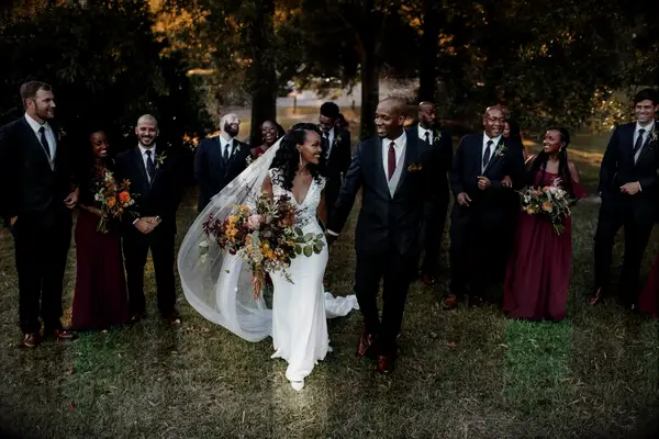 A bride and groom walk towards the camera, surrounded by their beaming wedding party.