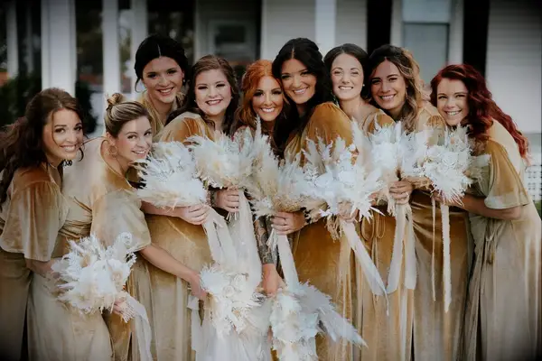 A bride poses for a photo with her wedding party, each of whom where autumnal-gold dresses and carry pampas grass bouquets.