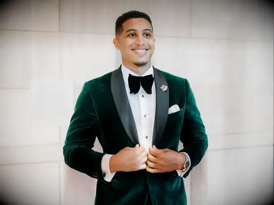 A groom smiles, wearing a vibrant autumnal-green velvet tuxedo jacket.