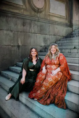 A couple pose on the steps of their venue, one bride wearing a jewel-toned green gown, the other glowing in an autumn-coral colored dress.