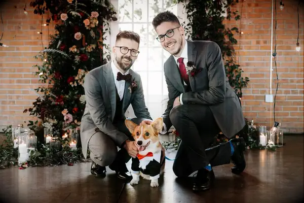 A pair of grooms lean down over their pup of honor, a little corgi in his own doggie-sized suit and tie.
