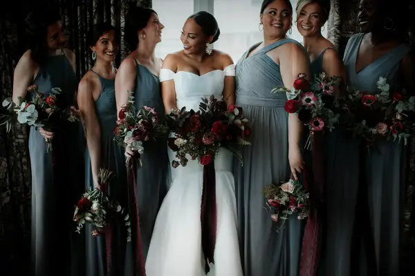 bride stands with bridesmaids wearing muted green chiffon dresses and holding bouquets with dark red flowers