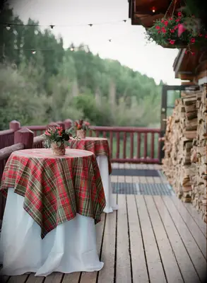 tall cocktail tables decorated with red and green plaid tablecloths arranged on wooden deck at outdoor rustic cabin wedding venue with mountain view in the background