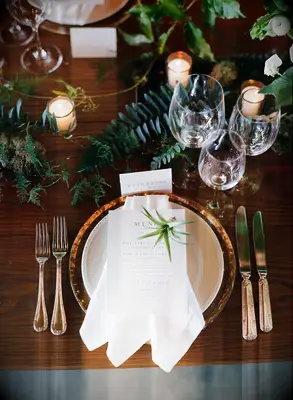 winter wedding place setting with gold charger plate, white folded napkin, votive candles and greenery against a bare wooden table