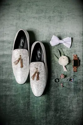 wedding photography flatlay with silver embroidered loafers decorated with gold tassels against green velvet backdrop. an apple watch, white bow tie and ranunculus boutonniere are placed next to the shoes