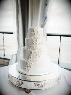 four-tier white fondant wedding cake with 3D sugar flower decorations on top of round silver cake stand