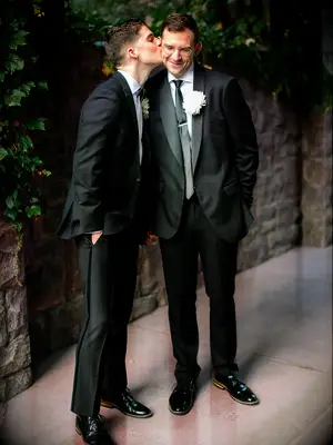 two grooms standing in garden wearing black tuxedos one groom kisses the other