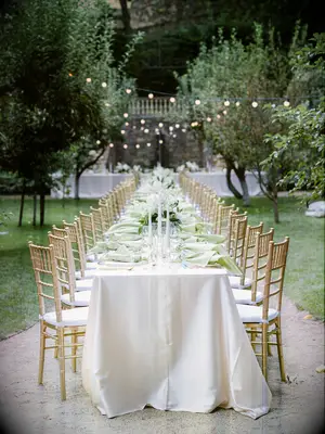 outdoor wedding reception with long banquet table decorated with iridescent cream table linens, taper candles and gold chiavari chairs