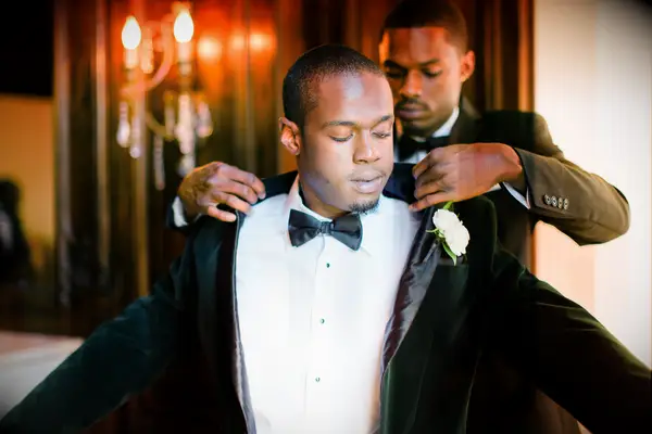Groomsman putting a green velvet blazer on the groom.